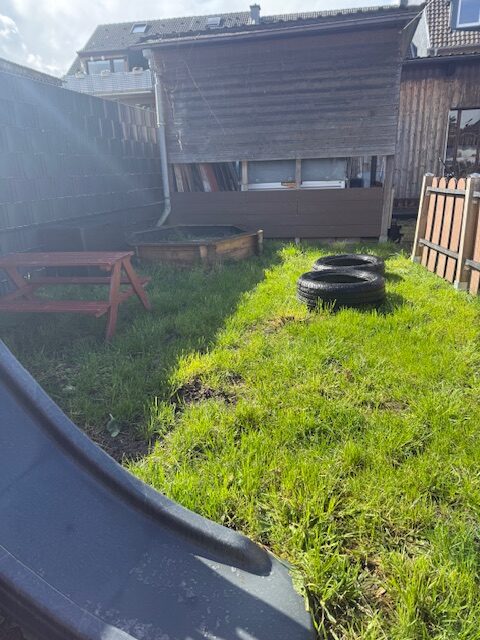 Der eingezäunte Spielbereich mit Picpic Bank und Sandkasten sowie Reifen zum klettern